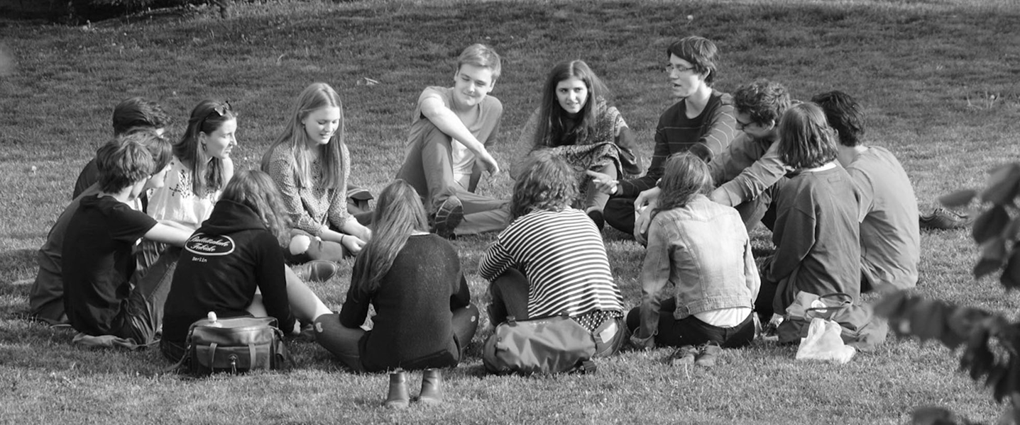 Grupa młodych ludzi siedzi w kręgu na trawie podczas rozmowy.